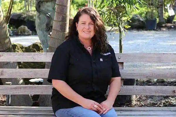 Woman with wavy hair sits on a wooden bench outdoors, wearing a black shirt and jeans, smiling. Background shows palm trees and greenery.