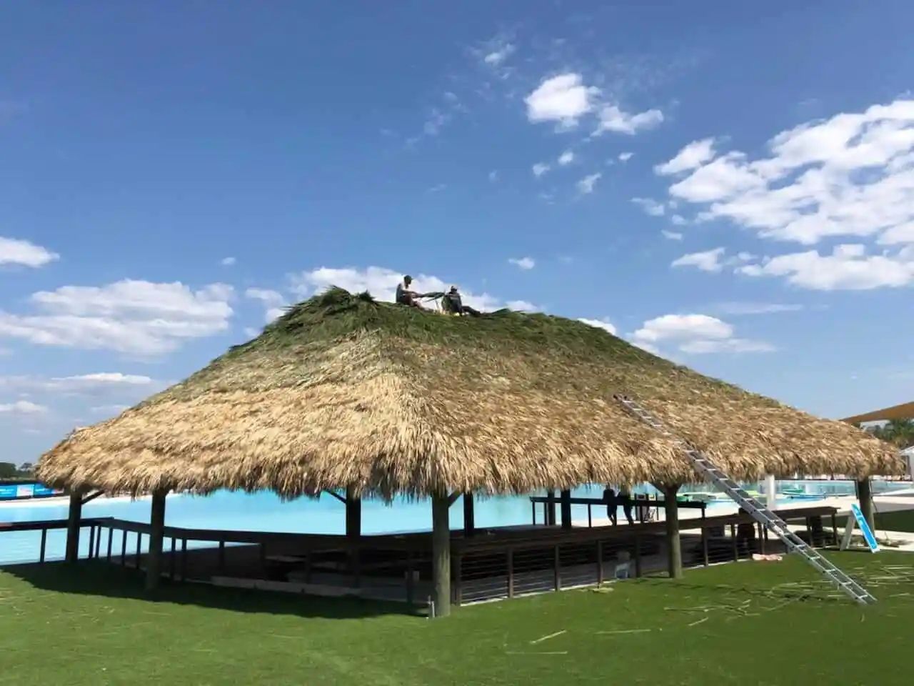 A large thatched-roof hut with a blue sky background, potentially over a pool or water feature. Two figures are working on the roof.