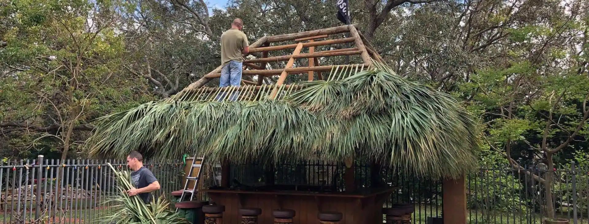 Two men are building a thatched roof structure. One man is on the roof, while the other stands below with materials.