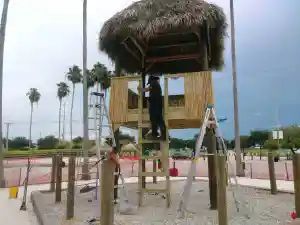 Construction workers building a wooden playhouse with a thatched roof in an outdoor park setting with palm trees.