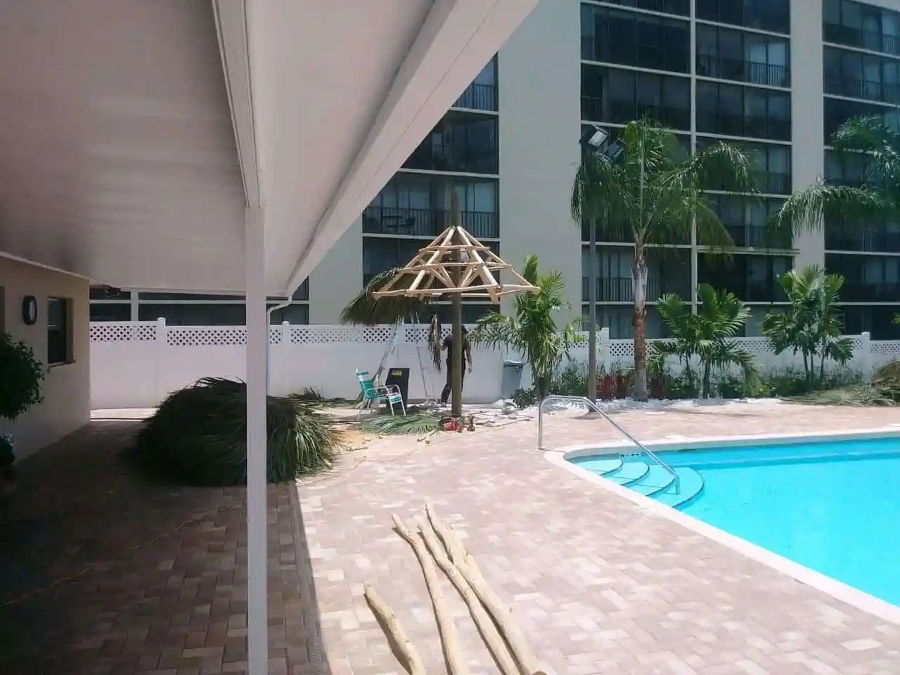 A covered patio with a pool, palm trees, and apartment building in the background; construction work on a wooden gazebo is underway.
