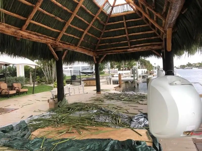 A thatched-roof gazebo by water, with cut greenery on the ground. A boat and dock are in the background.