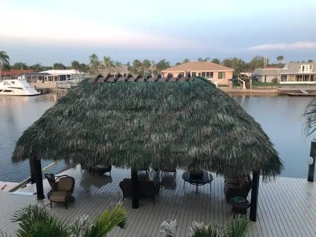 Tiki hut with thatched roof over a deck with seating, overlooking a waterway with boats and houses.