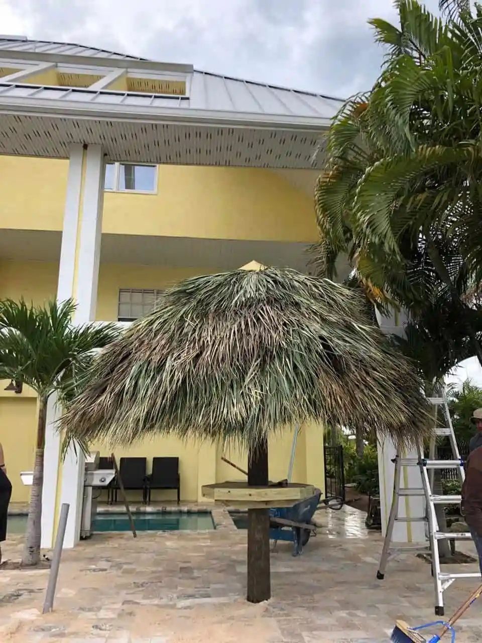 A thatched-roof umbrella near a yellow building with palm trees and a ladder, likely outdoors.