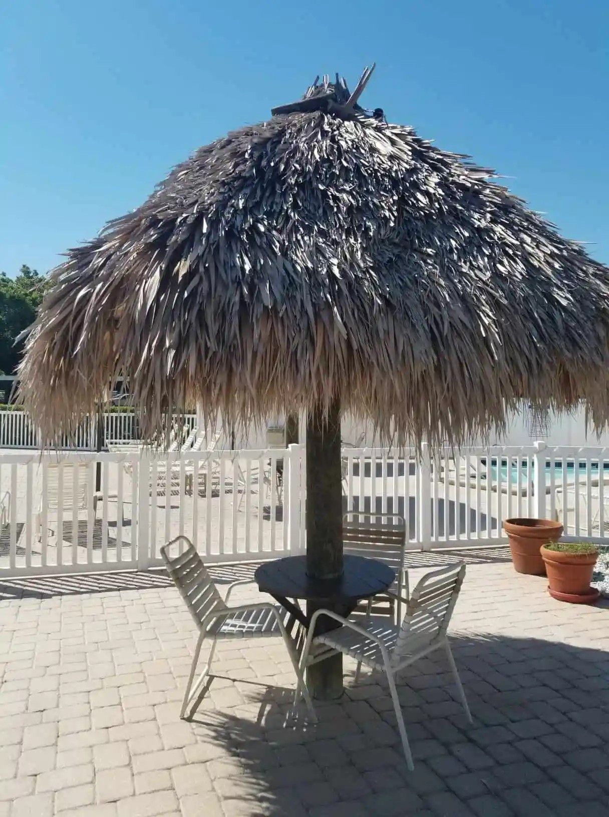 Tiki hut with a thatched roof over a table and two chairs on a brick patio next to a white fence and pool.