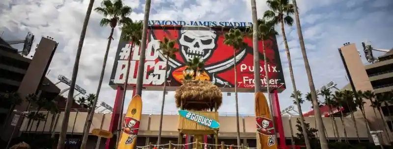 Raymond James Stadium with Buccaneers logo, palm trees, and tiki-themed structure in front.