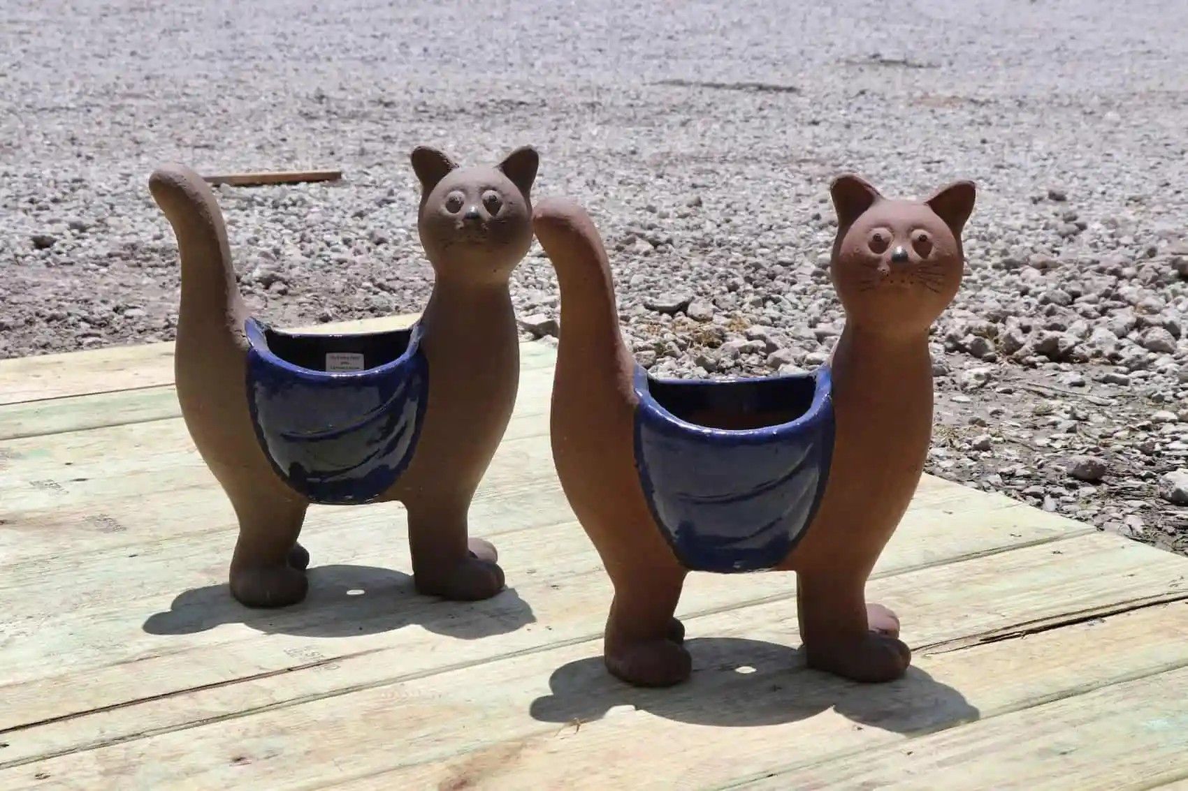 Two brown cat-shaped planters with blue pockets; sitting on wooden planks outdoors in sunlight.