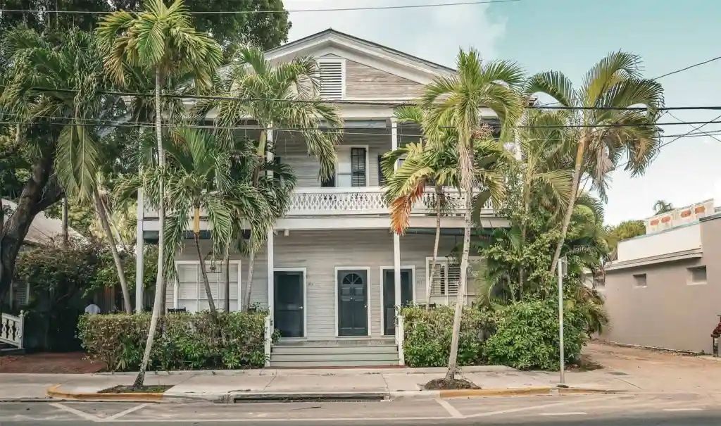 Two-story, pale grey house with a balcony, three dark doors below. Palm trees and shrubs line the front.
