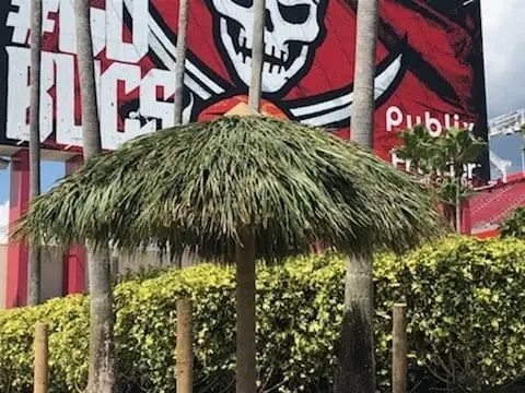 Tiki umbrella with a thatched roof in front of a Tampa Bay Buccaneers sign and palm trees.