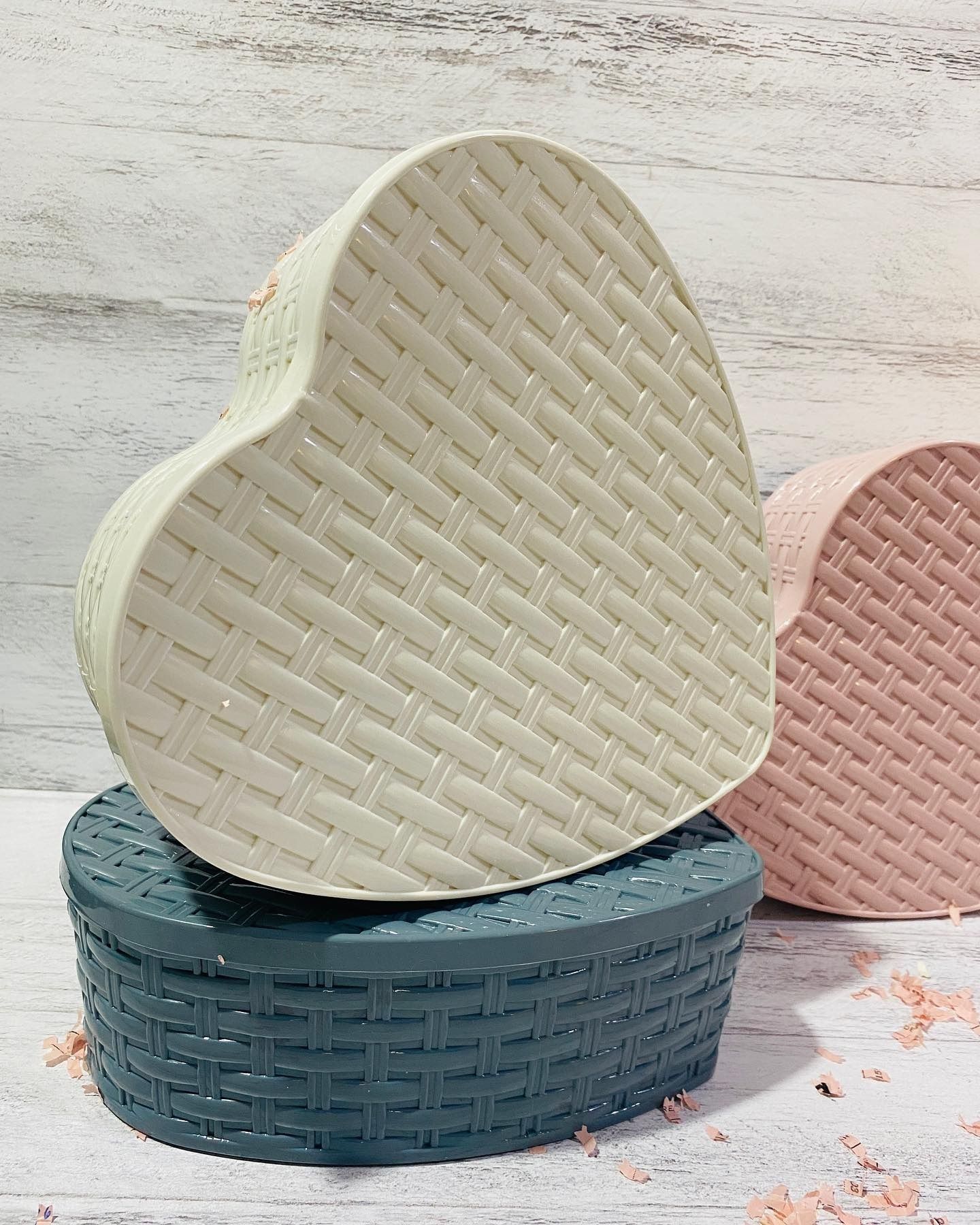 Tres cajas tejidas en forma de corazón: de color blanquecino, rosa y verde azulado, sobre un fondo de madera clara.