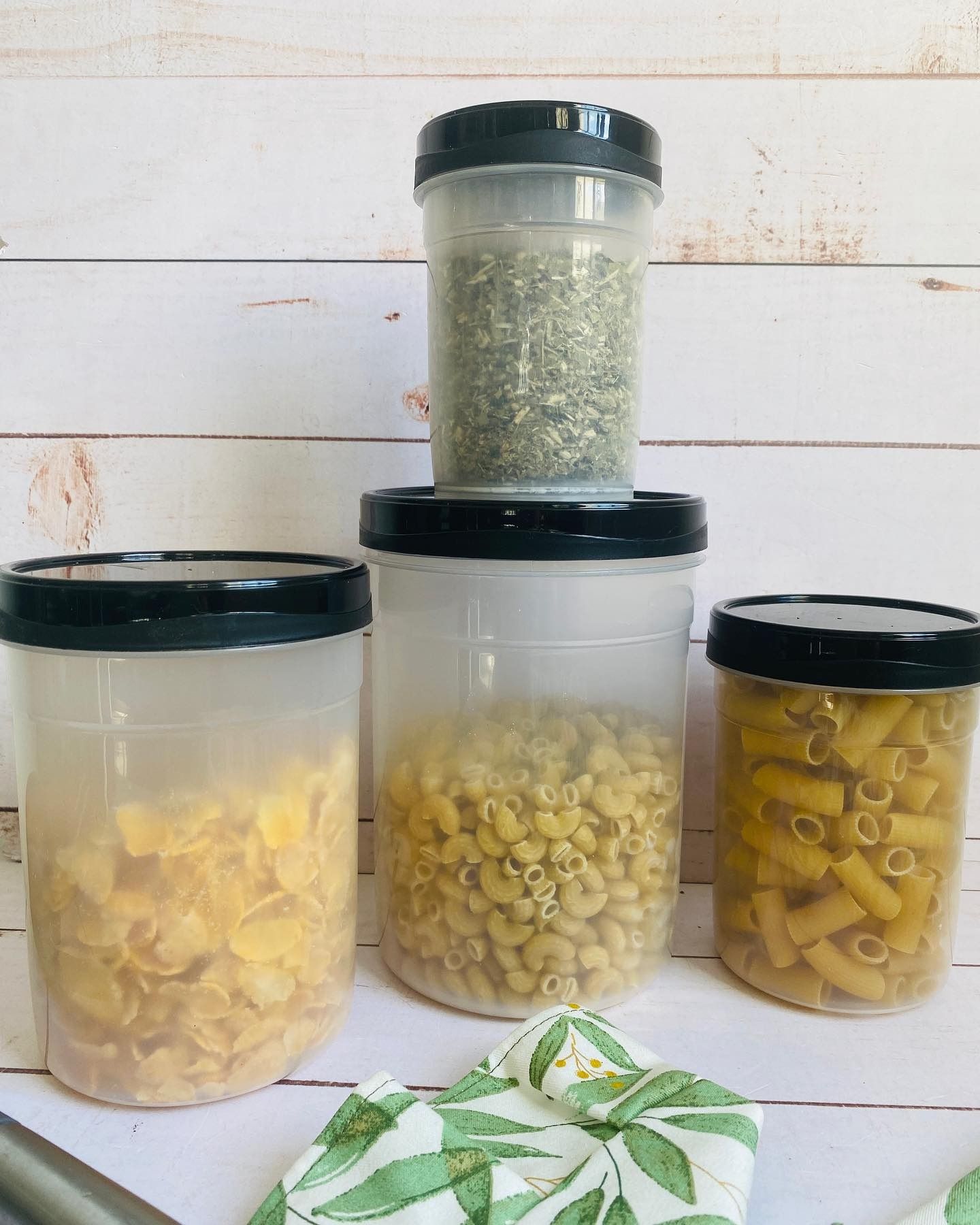 Pasta y hierbas en recipientes de plástico transparente y herméticos con tapas negras. Sobre un fondo de madera blanca.