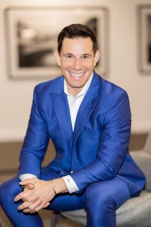 A man in a blue suit is sitting on a chair with his hands folded.