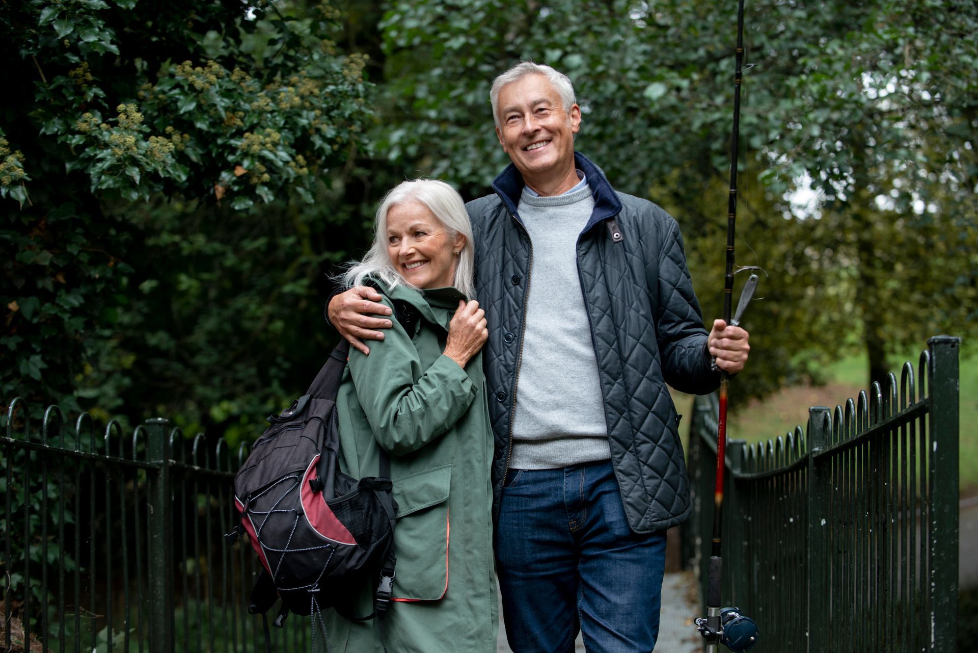 A man and a woman are standing next to each other in a park . the man is holding a fishing rod.