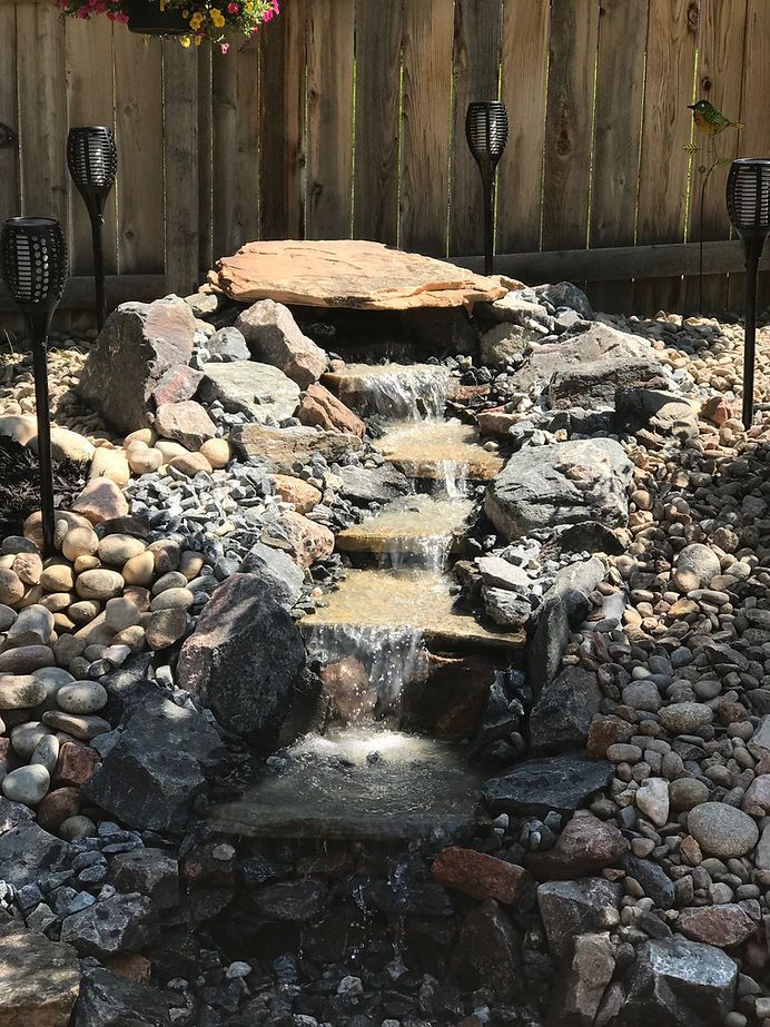 A small waterfall is surrounded by rocks and a wooden fence.