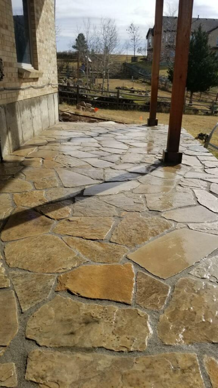 A stone patio with a wooden pergola and a house in the background.