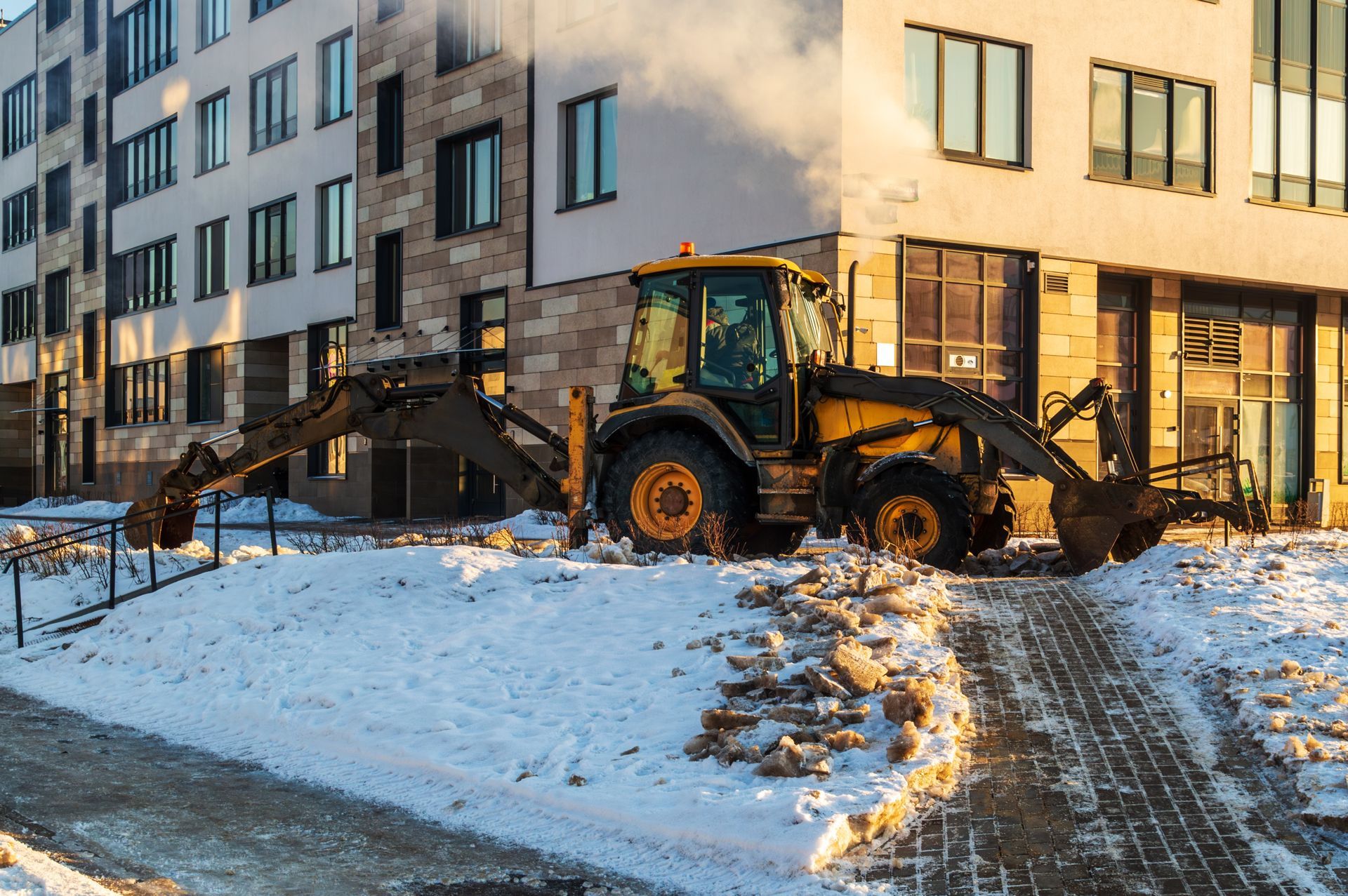 Yellow backhoe on a snow-covered construction site next to a modern building.