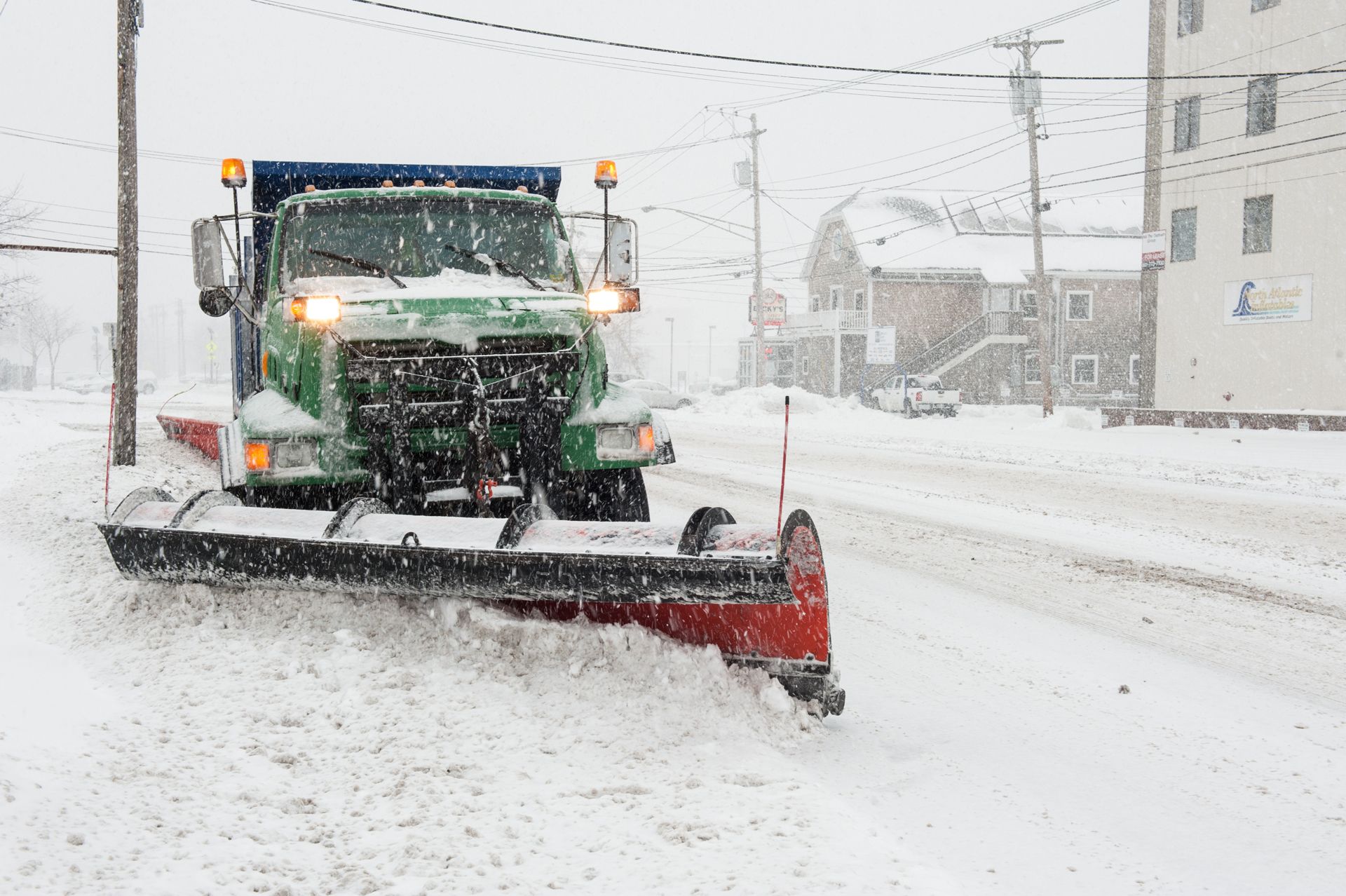 Snow plow clearing a snow-covered street during a snowfall; green truck with orange lights.