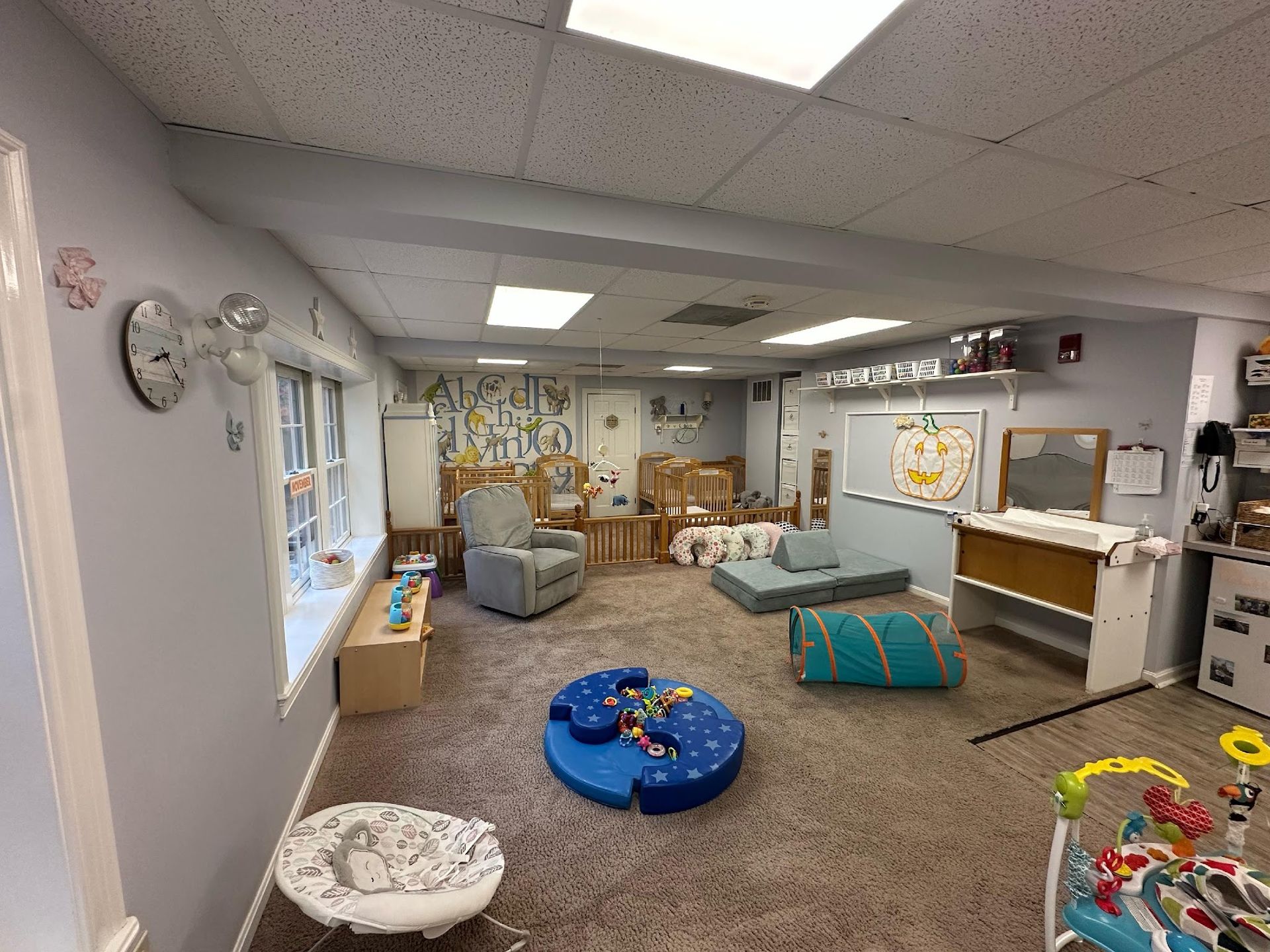 A brightly lit childcare room with floor cushions, a blue play tunnel, baby bouncers, and cribs on a carpeted floor.