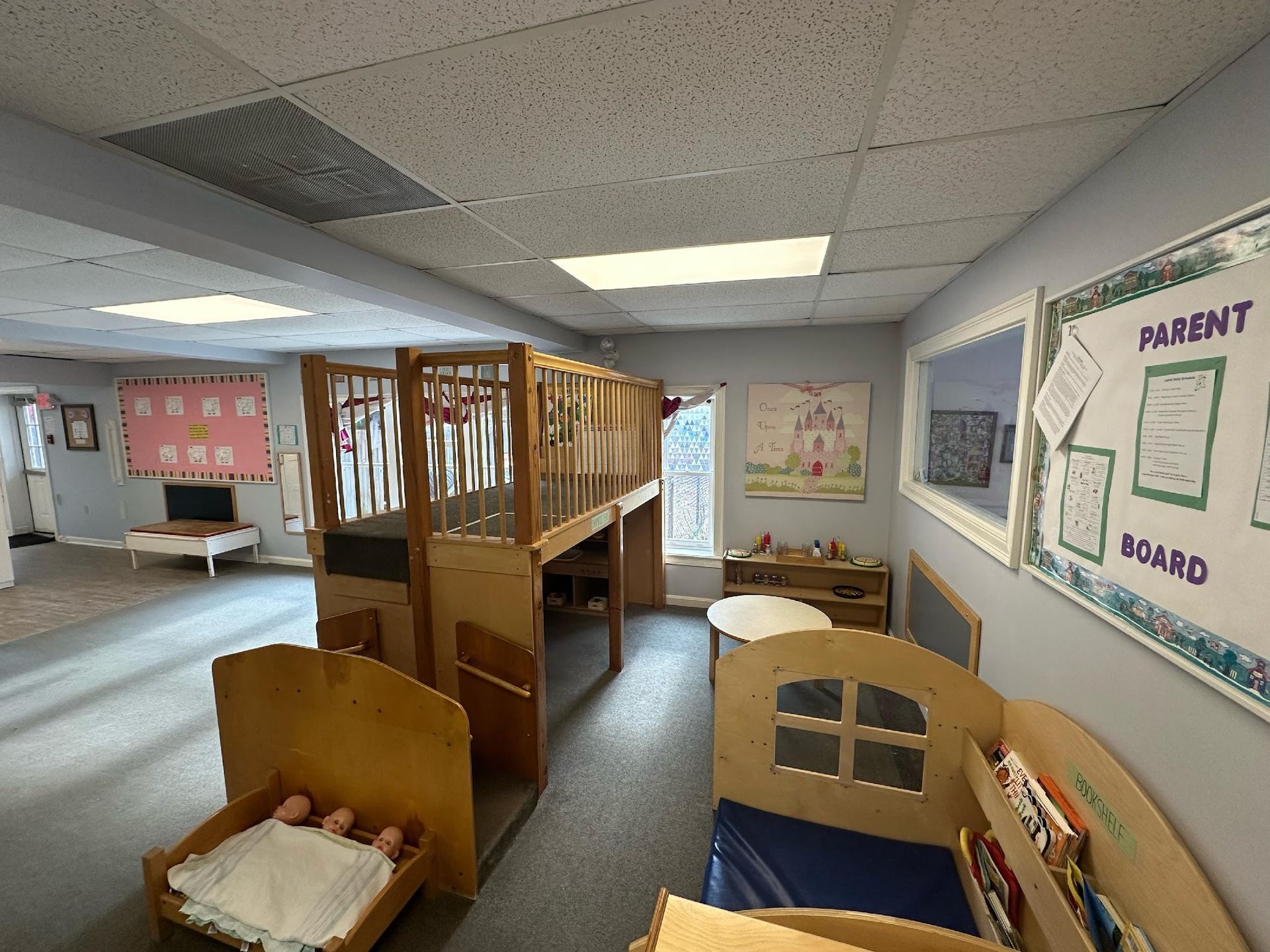 A brightly lit preschool classroom featuring a wooden loft structure, a small toy bed, and a parent information board.