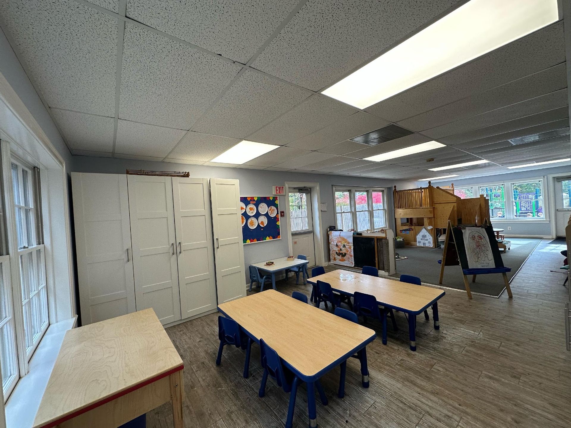 A brightly lit classroom featuring wooden tables, small blue chairs, white storage cabinets, and a play area in the back.