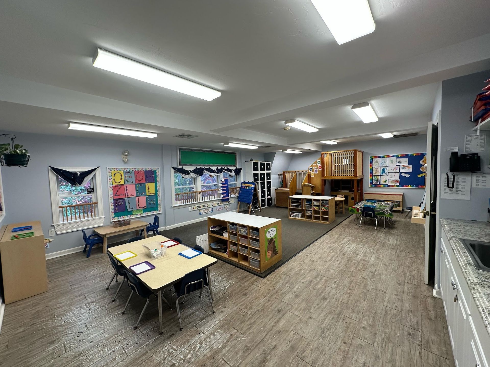A brightly lit preschool classroom with tables, a wooden play structure, toy storage shelves, and learning wall displays.
