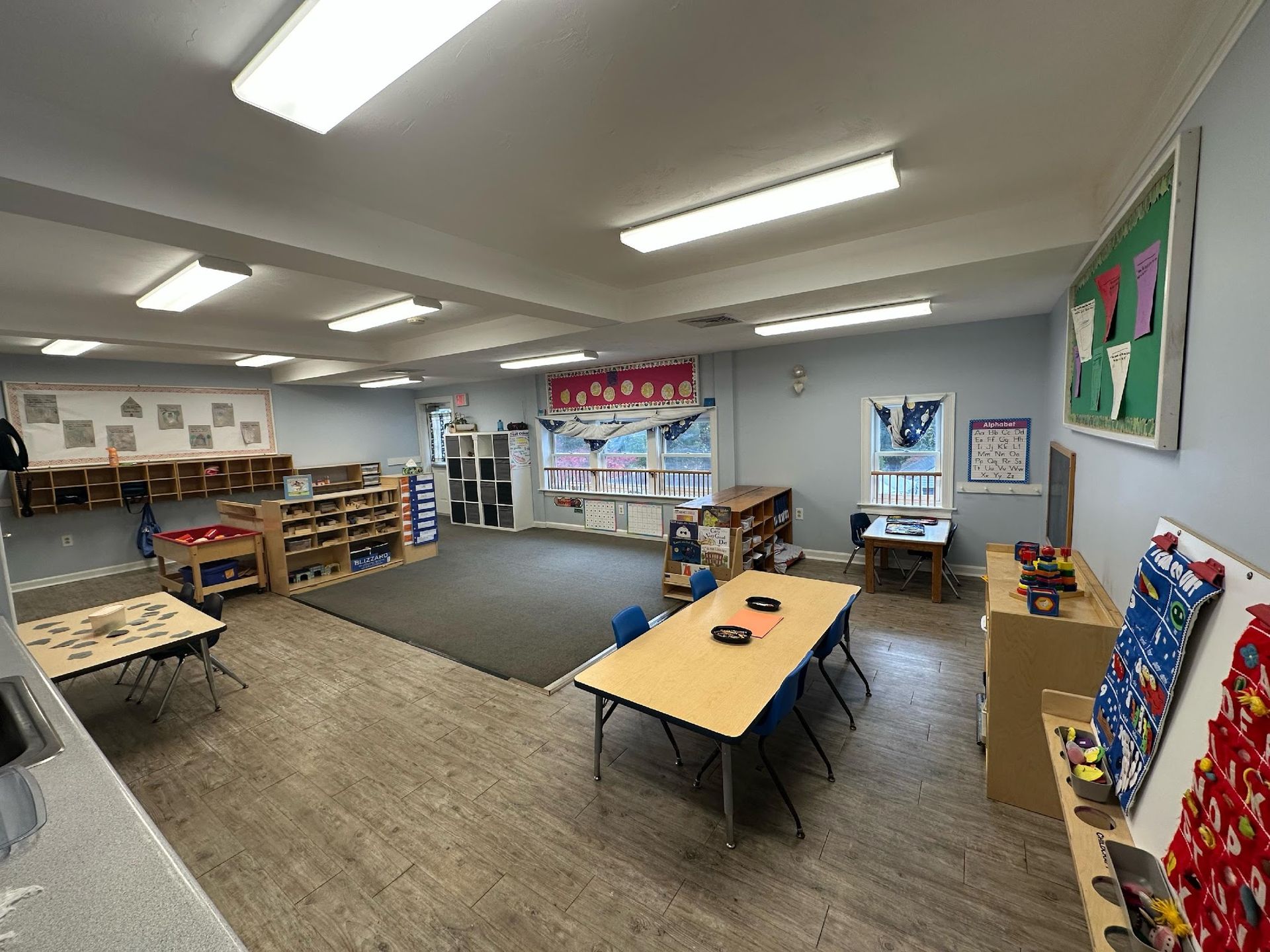 An empty preschool classroom with tables, chairs, shelving units, and educational posters on light blue walls.