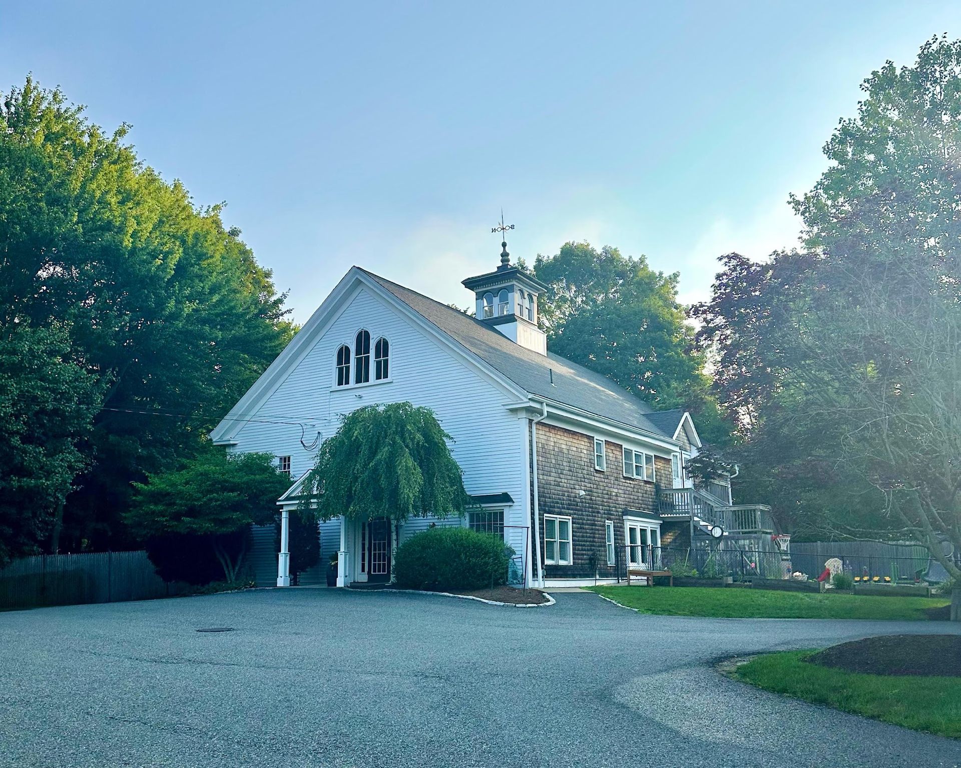 A white, multi-level house with a cupola and arched windows, surrounded by green trees over a gravel driveway.