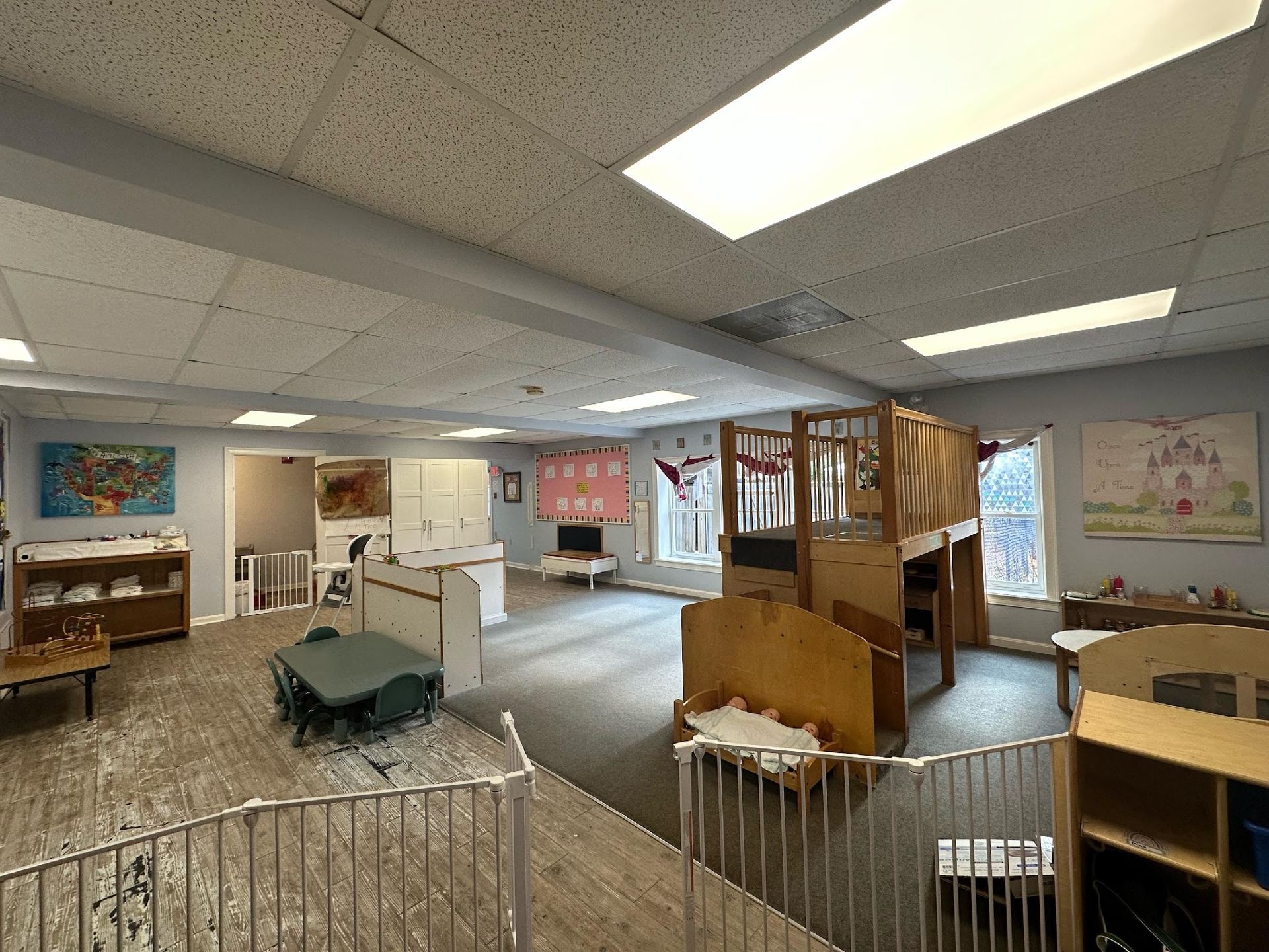 A bright, spacious indoor daycare room featuring a wooden loft play structure, children's tables, and colorful wall art.