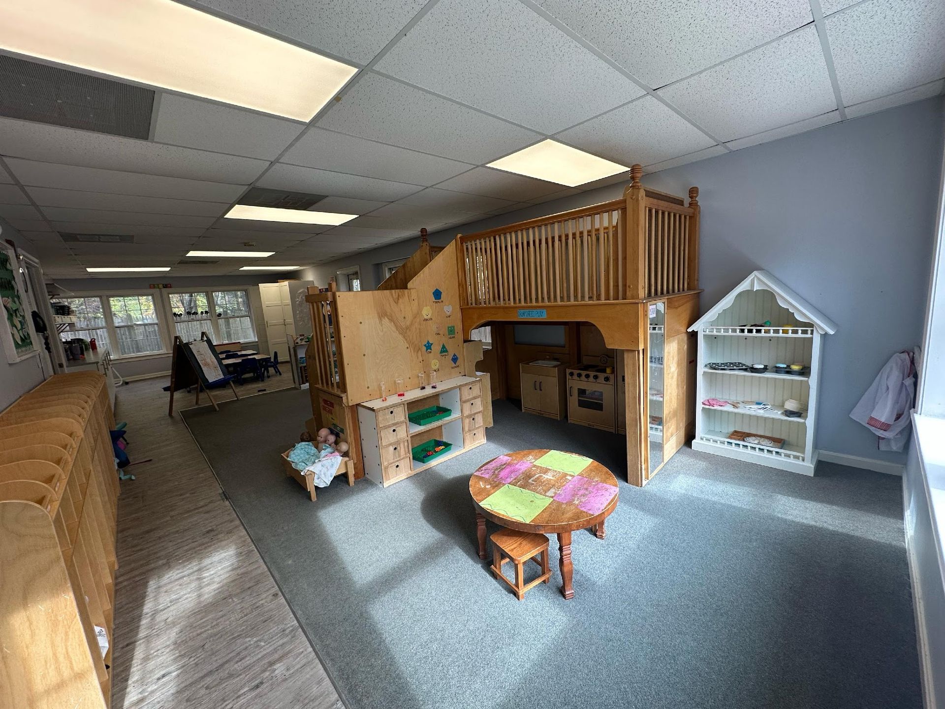 A bright preschool classroom with a wooden loft, a small circular table, chairs, and a white house-shaped bookshelf.