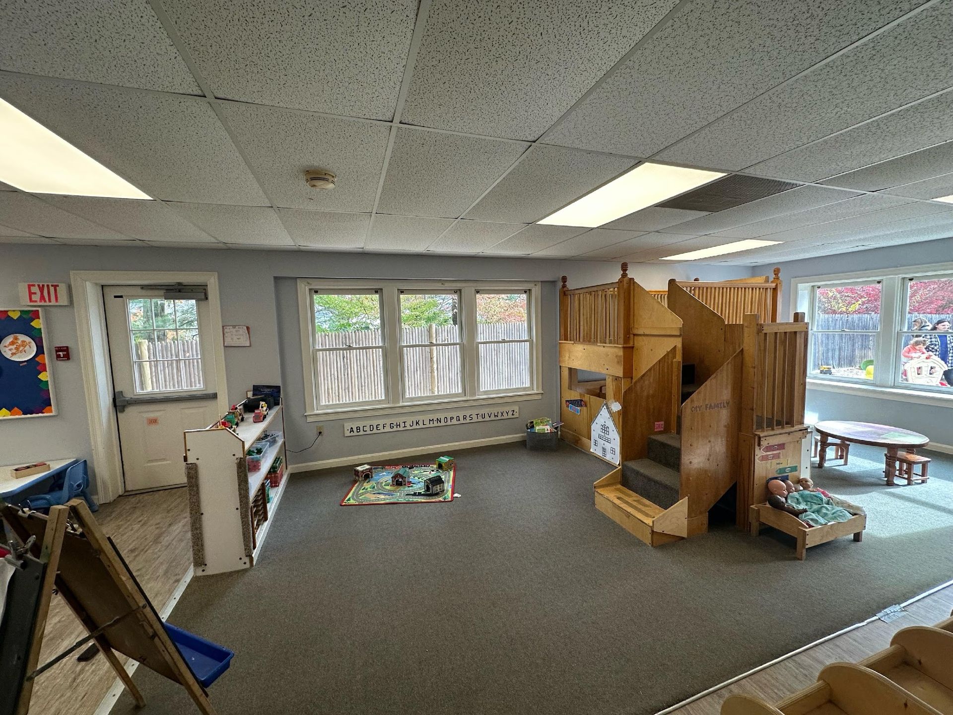 A brightly lit preschool classroom with gray carpet, large windows, a wooden play loft, and various toys and furniture.