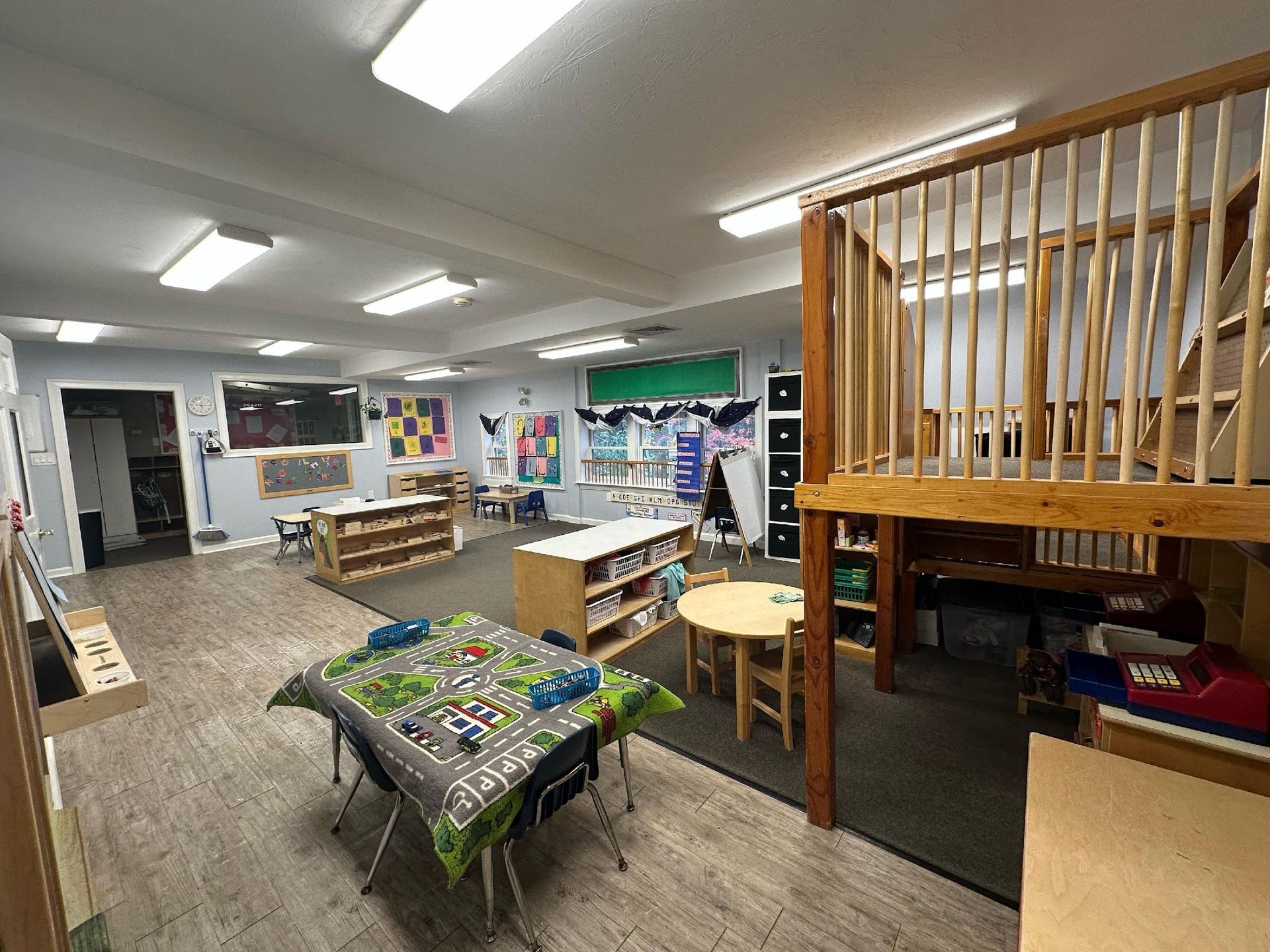 A bright classroom for young children featuring a wooden loft play area, small tables, and toy storage shelves.