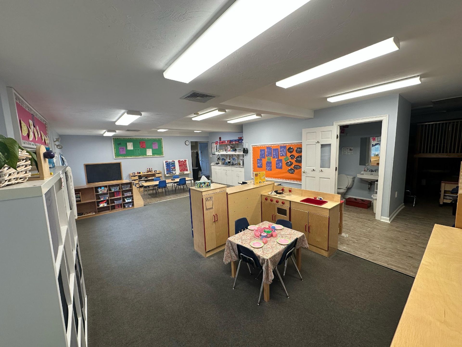 A preschool classroom featuring a play kitchen, a small table with chairs, shelves, and educational posters on blue walls.