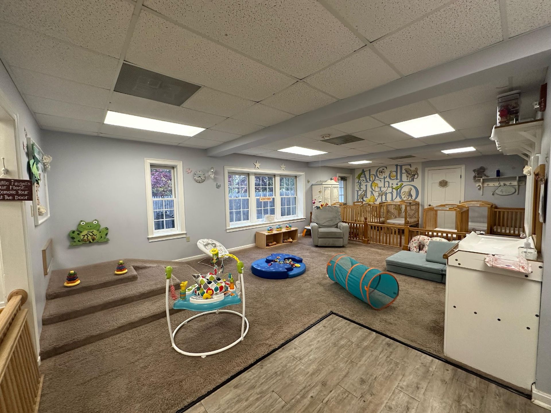 A brightly lit daycare room with carpeted floor, infant activity gear, a play tunnel, and cribs lined up against the wall.