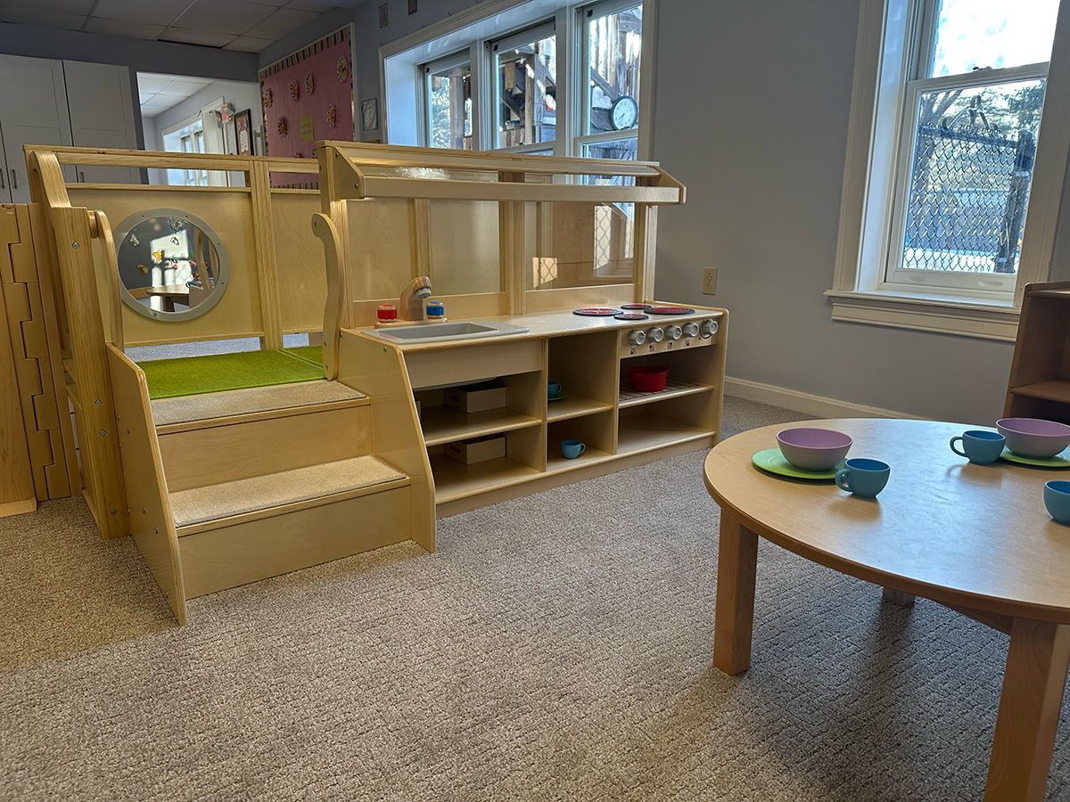A light wood toddler kitchen playset with steps sits in a carpeted classroom near a round table with toy tea sets.