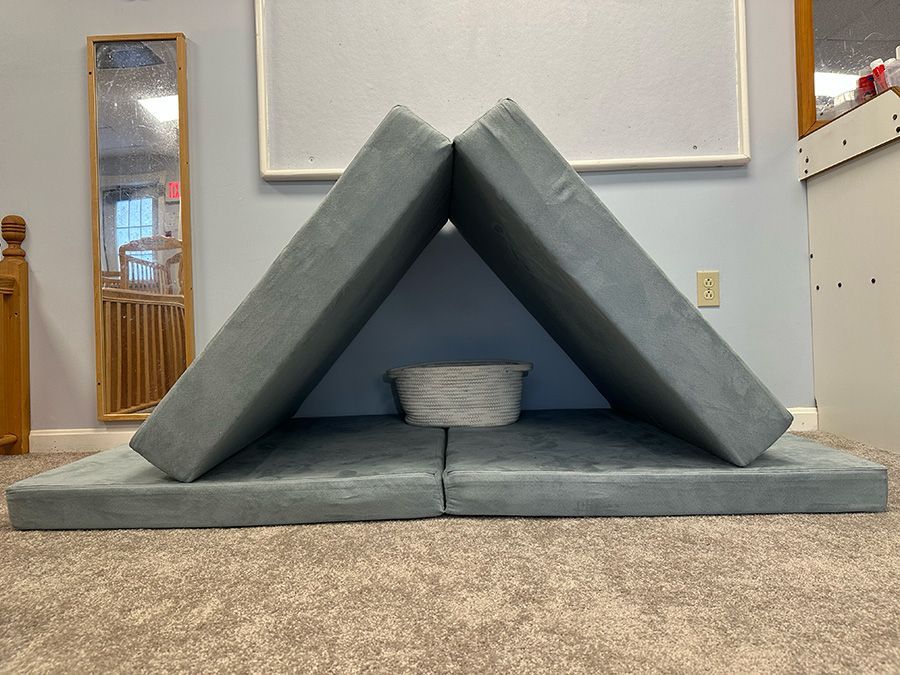 A grey foam play mat folded into a triangle tent shape against a wall, with a small white basket inside.