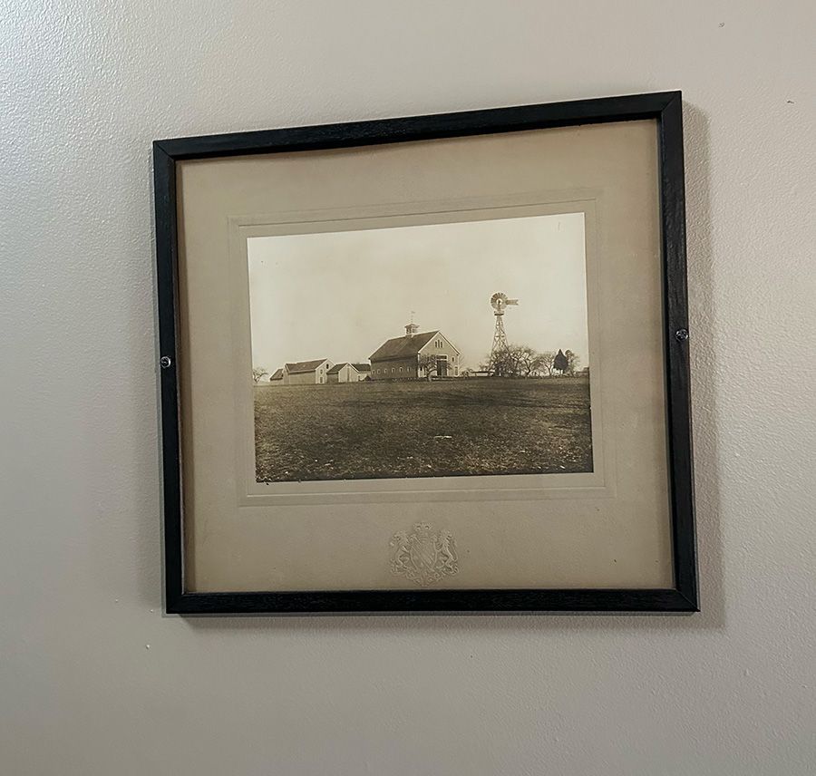 A framed, sepia-toned vintage photograph of a rural farmhouse and windmill in a flat, open field.