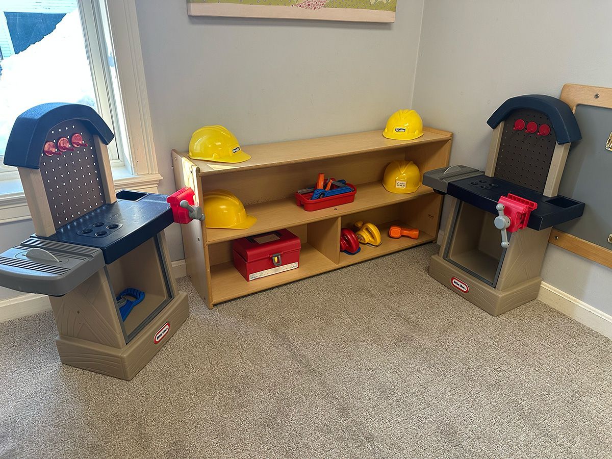 Two toy workbenches and a wooden shelf stocked with yellow construction hats and red tool accessories on a carpeted floor.