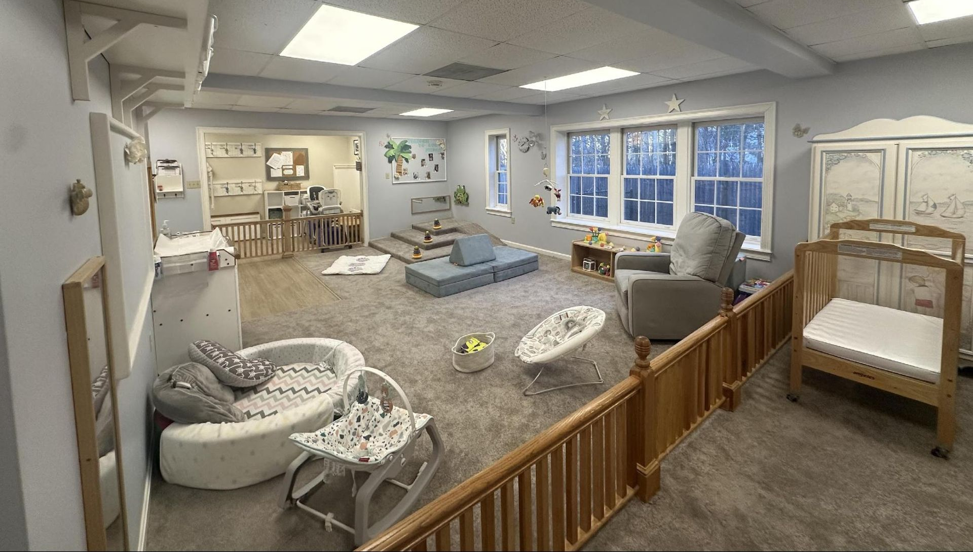 A bright infant childcare room with a crib, play mats, bouncers, and a soft-toned sitting area with carpeted floors.