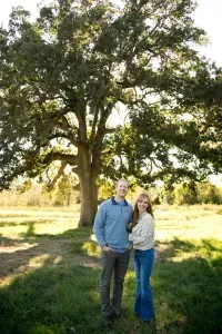 A man and a woman are standing in front of a large tree in a field.