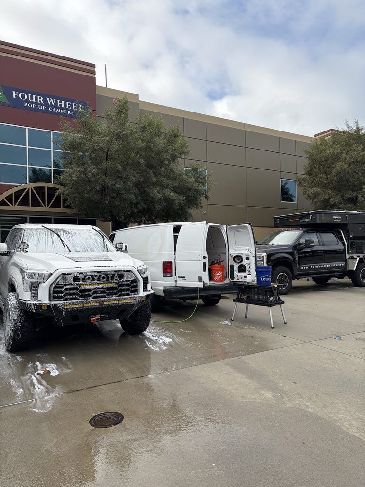 Three vehicles being washed in front of a building: white truck, white van, and black truck.