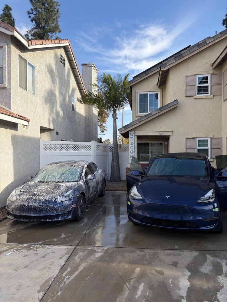 Two blue Tesla cars parked in front of townhouses. One car is covered in soap suds. Sunny day.