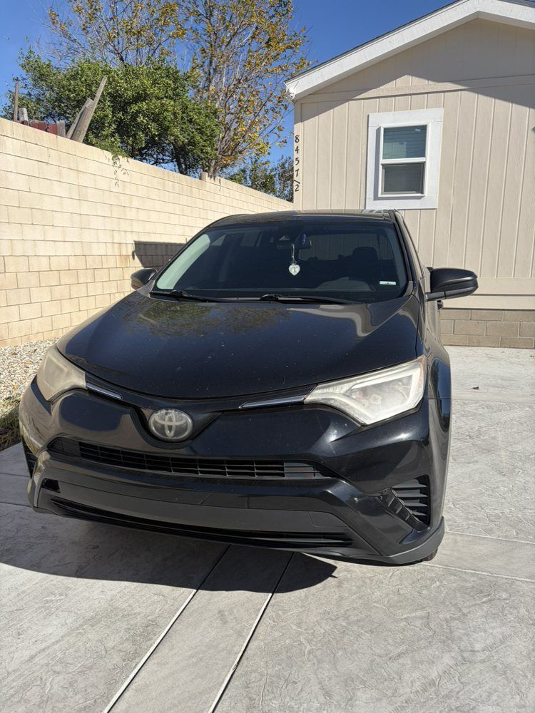 Black Toyota RAV4 parked in front of a beige house with a brick wall on a sunny day.