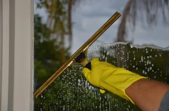 Hand in yellow glove uses a squeegee to clean a soapy window.