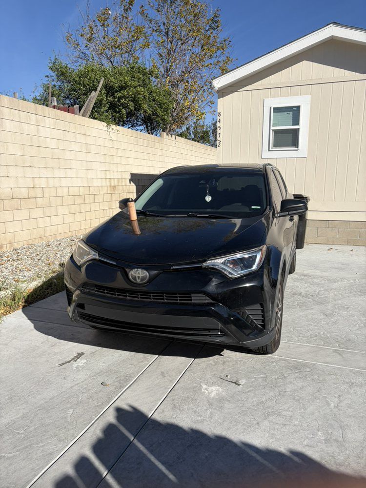 Black Toyota RAV4 parked in a concrete driveway near a beige house and brick wall.