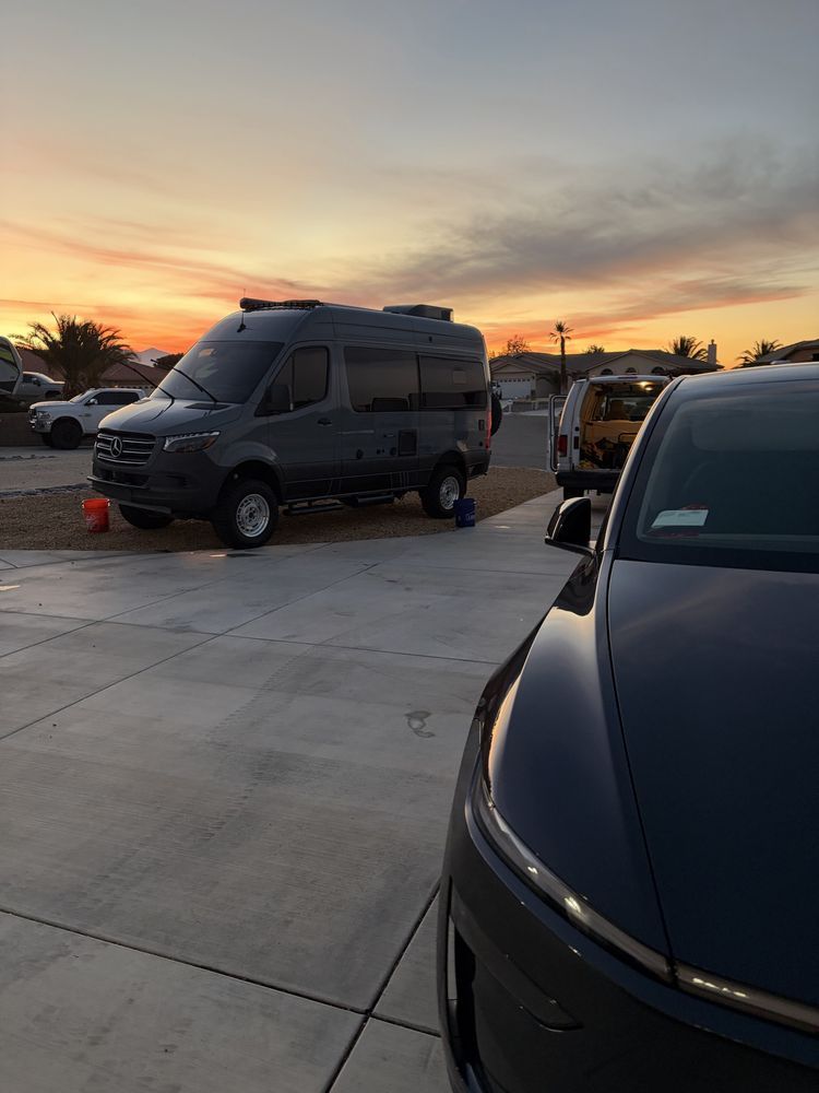 A dark gray camper van and black car parked on a paved lot at sunset.