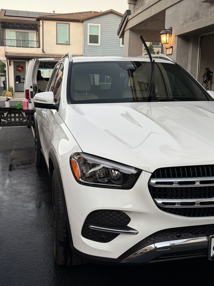 White Mercedes SUV being washed in a driveway, house in the background. Windshield wipers up.