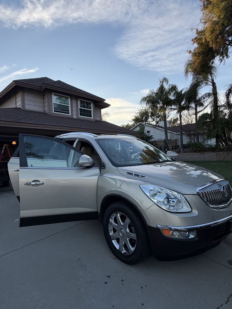 Tan Buick Enclave SUV parked in a driveway with open doors; house and trees in the background under a cloudy sky.