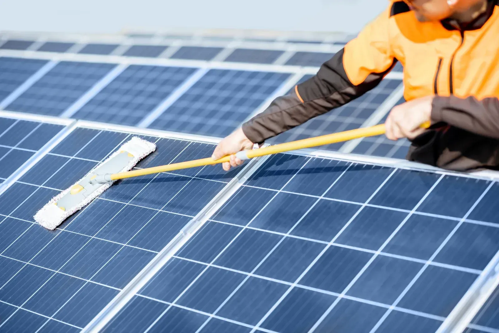 Person in orange jacket cleaning solar panels with a long-handled brush on a sunny day.