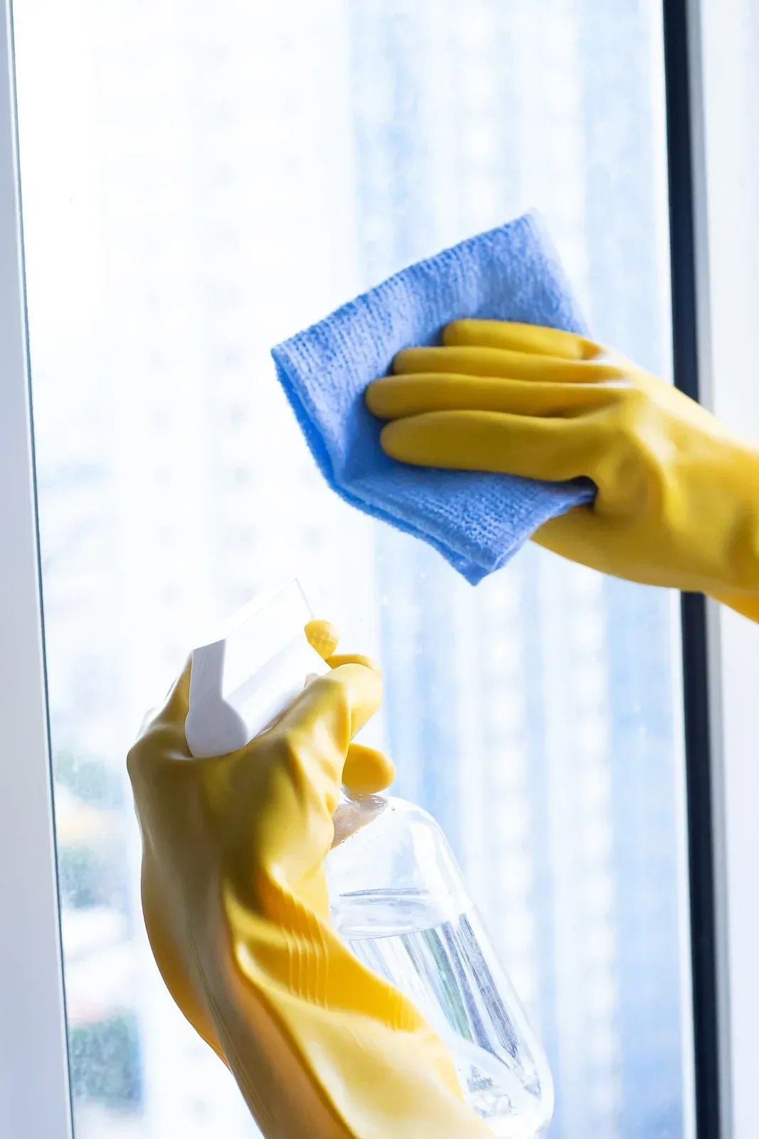 Person wearing yellow gloves spraying and wiping a window with a blue cloth.