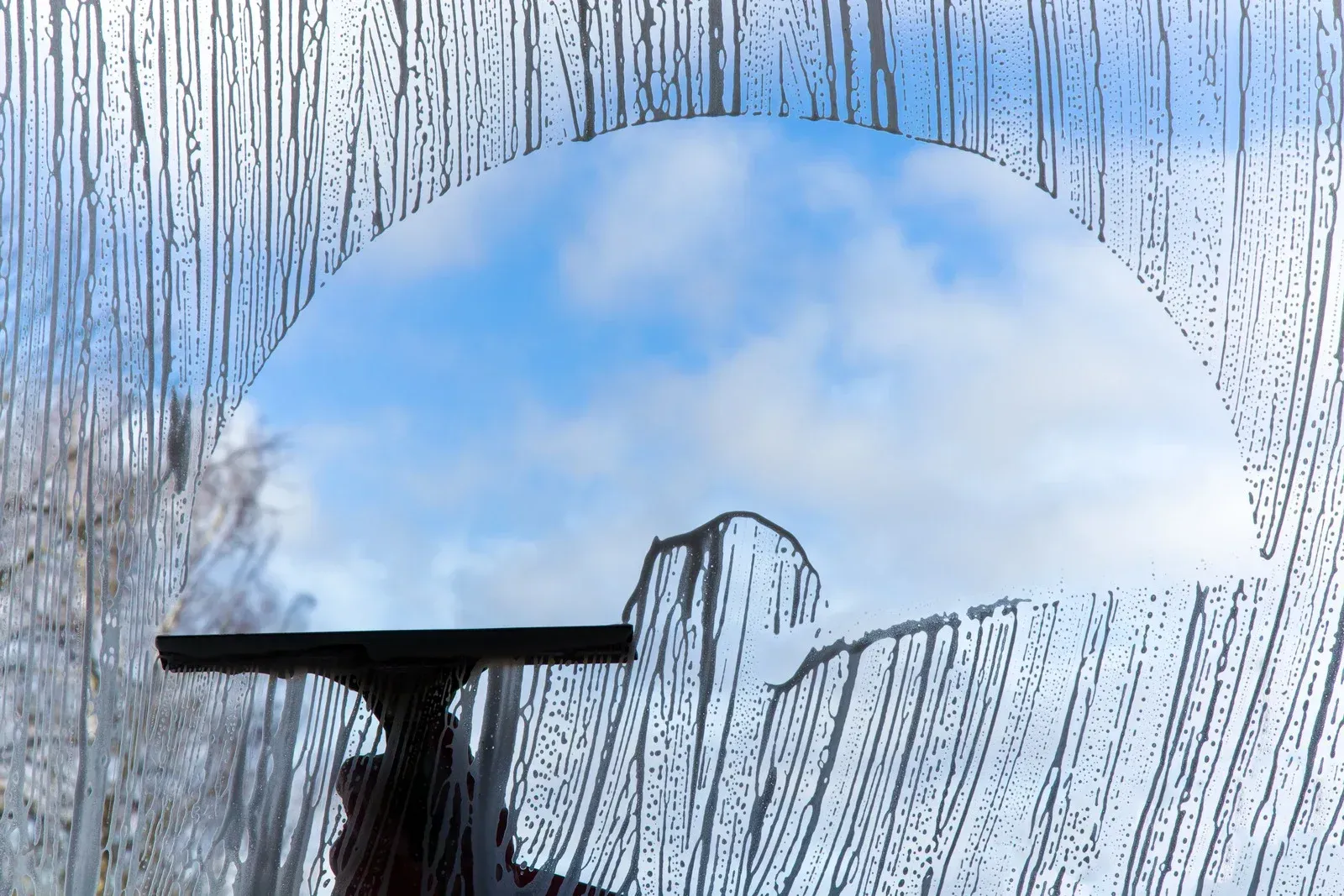 A hand squeegees a window, leaving a semicircular clear area revealing blue sky and clouds.